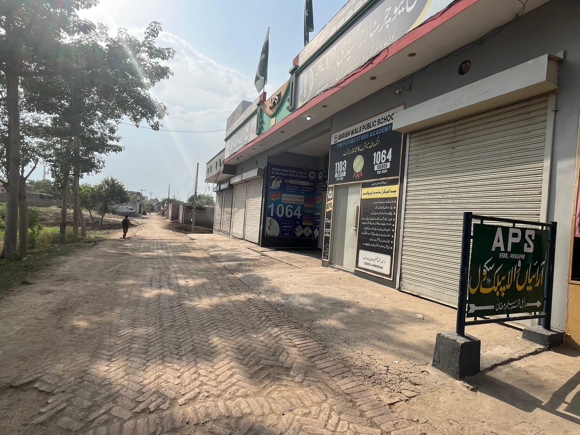 Arrian Wala Public School seen from the right, with the green APS gate sign in the foreground and the school entrance and signboards along the street behind it.